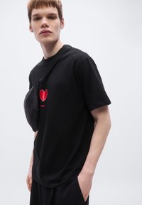 Μαύρο βαμβακερό μπλουζάκι με κόκκινη καρδιά στο στήθος. Το μοντέλο φοράει μαύρο τσαντάκι μέσης περασμένο στον ώμο.