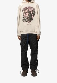 Sudadera crema con un diseño gráfico de león, combinada con pantalones cargo negros que cuentan con múltiples bolsillos y un acabado texturizado. Zapatillas marrones.