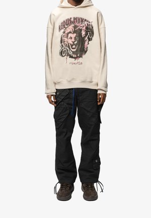 Sudadera crema con un diseño gráfico de león, combinada con pantalones cargo negros que cuentan con múltiples bolsillos y un acabado texturizado. Zapatillas marrones.