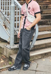 Osoba opřená o kovový zábradlí blízko betonových schodů, oblečená v růžovém a černém sportovním dresu, černých džínách, teniskách Nike a s křížovou taškou.