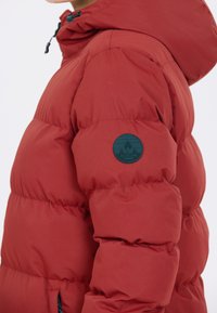 Rød pufferjakke med hætte, der har et tekstureret stof, vandret syninger og et rundt logo-patch på venstre ærme.