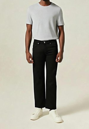 Hombre con camiseta de manga corta gris claro, pantalones negros de pierna recta y zapatillas blancas, de pie frente a un fondo liso.