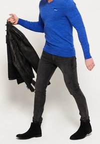 Blauer long-sleeve V-Ausschnitt Pullover, schwarze lässige Slim-Fit Jeans und schwarze Wildleder-Stiefeletten, hält eine schwarze Lederjacke.