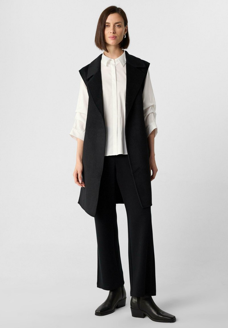 Schwarze ärmellose Weste mit Reverskragen, getragen über einer weißen Bluse. Das Modell kombiniert sie mit schwarzen Hosen und Stiefeletten, was ein minimalistisches Design zeigt.