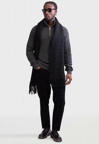 Camisola cinza escura com fecho de correr, textura canelada, cachecol preto com franjas, calças pretas e sapatos pretos. Acessórios incluem óculos de sol.