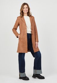 Brauner Leder-Trenchcoat mit Kragen, einreihigen Knöpfen und Seitentaschen. Kombiniert mit dunkelblauen weit geschnittenen Jeans und schwarzen Stiefeln.