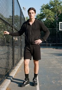 Jeune homme en sweat à capuche noir, short et baskets, debout sur un terrain de basketball en plein air à côté d'une clôture en chaîne avec une main sur la hanche.