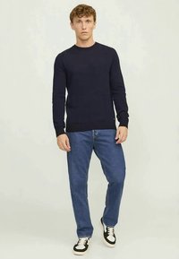 Man wearing a black long-sleeve sweater, blue jeans, and black-and-white sneakers standing against a plain light background.