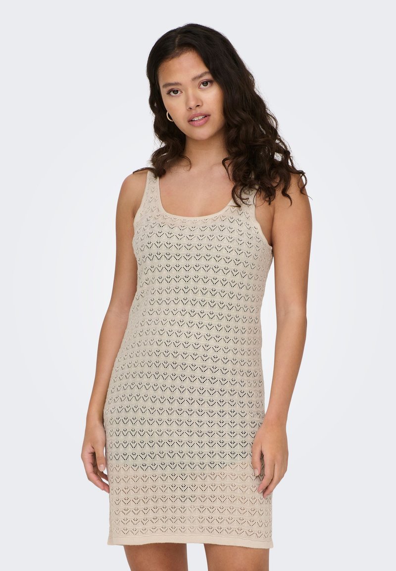 Jeune femme aux longs cheveux bouclés, portant une robe beige sans manches en crochet, debout devant un fond uni gris clair.