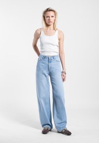 Lichtblauwe, high-waisted jeans met wijde pijpen, gemaakt van denim. Gecombineerd met een witte geribbelde tanktop. Het model staat tegen een effen achtergrond.