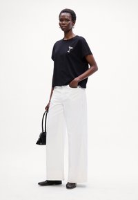 Sort t-shirt med et drikke-grafik, parret med hvide brede bukser. Modellen holder en lille sort håndtaske og har sorte sko på.