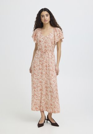 Naine, kellel on pikad lokkis juuksed, kannab beežikat lillelist midi-kleit lillelise varrukaga ja musta terava ninaosaga kontsakingi, seisab valge tausta ees.