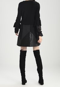 Pull en maille noir à manches volantes, associé à une jupe en cuir noir avec un motif géométrique, et des bottes en daim noir jusqu'au genou.