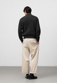 Maglione a coste verde scuro abbinato a pantaloni larghi color crema e scarpe nere, visto da dietro. Linee pulite e design semplice.