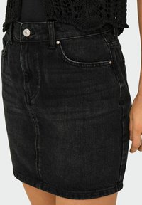 Zwarte denimrok met een getailleerde vorm, voorkant zakken, zilveren knoopsluiting en contrasterende stiksels aan de onderrand. Textuur afgewerkte stof.