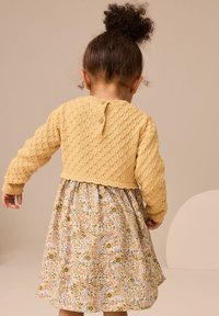 Pull en laine de couleur moutarde avec un motif texturé, associé à une robe fleurie aux couleurs pastel et à la coupe décontractée, froncée à la taille.