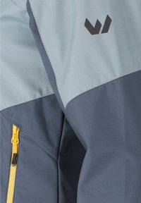 Gros plan d'une veste grise bicolore avec une fermeture éclair jaune et un logo géométrique noir sur le haut de la manche.