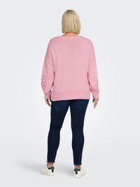 Pink oversized sweater with ribbed hem and cuffs, featuring a decorative design on the left sleeve, paired with dark skinny jeans and sneakers.
