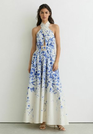 Vrouw leunend tegen een witte muur, gekleed in een witte halternek maxi-jurk met een blauw bloemenpatroon en een riem, gecombineerd met witte sandalen.