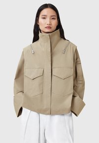 Giacca beige tagliata con colletto alto, due tasche anteriori e dettagli con zip sulle spalle. Realizzata in un tessuto liscio e leggero.