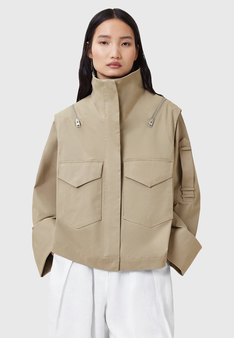 Giacca beige tagliata con colletto alto, due tasche anteriori e dettagli con zip sulle spalle. Realizzata in un tessuto liscio e leggero.