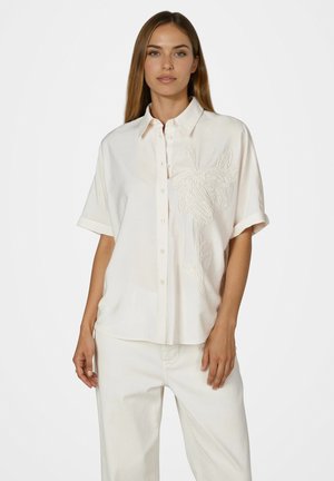 Frau trägt ein lockeres weißes Hemd mit kurzen Ärmeln und Blumenstickerei auf der Brust, kombiniert mit weißen Hosen.