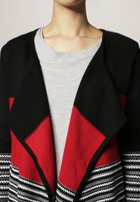 Cardigan nero con motivo geometrico rosso e bianco, caratterizzato da un davanti drappeggiato e una maglietta grigia sotto. Materiale in maglia testurizzata.