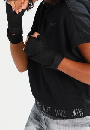 Osoba nosząca czarne opaski na nadgarstki i czarny top Nike, z rękami ustawionymi w pozycji bokserskiej lub walki.