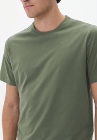 Tricou de culoare verde măsliniu cu guler rotund, confecționat din bumbac moale, cu mâneci scurte și o textură netedă, fără ornamente. Croi lejer.