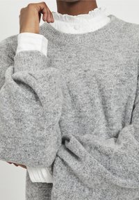Maglione grigio chiaro lavorato a maglia con maniche ampie, indossato sopra una camicetta bianca con colletto a frange e chiusura con bottoni. Materiale testurizzato visibile.