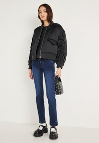 Chaqueta bomber de satén negra con cierre de cremallera y bolsillos laterales, combinada con jeans azul oscuro y zapatos de plataforma negros y voluminosos. Pequeño bolso tachonado.