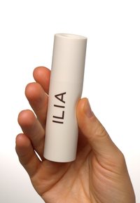 Contenitore cilindrico bianco con una parte superiore piatta, con testo scuro che recita "ILIA." Tenuto in una mano con un tono di pelle naturale. Finitura liscia e opaca.