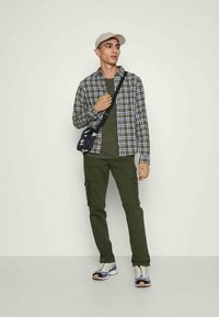 Jovem com um boné bege, camisa xadrez sobre uma t-shirt verde, calças cargo olive e ténis brancos, a carregar uma pequena bolsa preta de ombro.