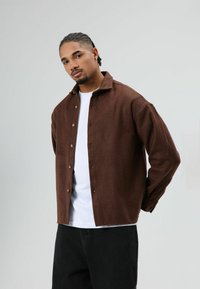 Bruin overhemd met knopen van zachte stof, voorzien van een klassieke kraag, knopen aan de voorkant en een ontspannen pasvorm, gedragen over een wit T-shirt.