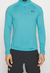 Türkises longsleeve Sportshirt mit einem halben Reißverschluss am Kragen und schwarzem Logo auf der Brust, gefertigt aus einem glatten, dehnbaren Material.