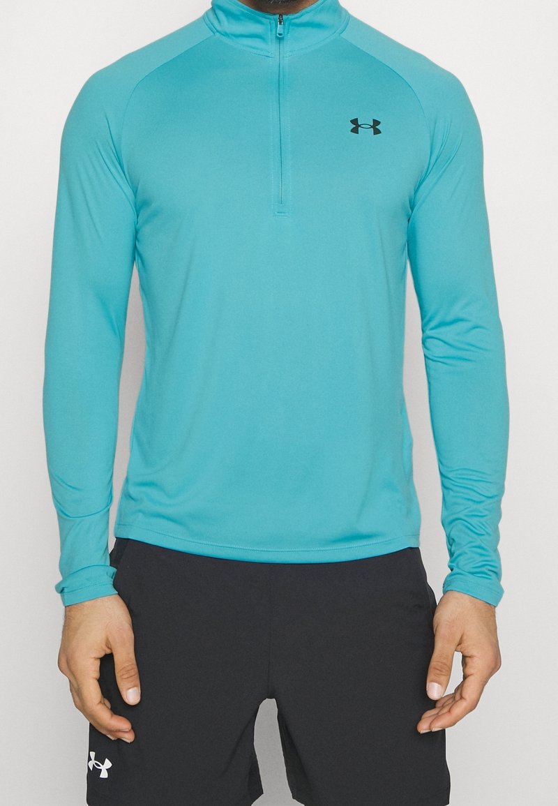 Türkises longsleeve Sportshirt mit einem halben Reißverschluss am Kragen und schwarzem Logo auf der Brust, gefertigt aus einem glatten, dehnbaren Material.