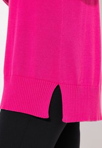 Fuchsia stickad tröja med ribbad nederkant och sidoflikar, med en slät yta och avslappnad passform, kombinerad med svarta leggings.