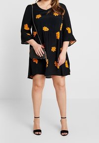 Mustat mekko, jossa on lyhyet kellohihat, ja siinä on oranssit kukkakuviot. Tyylitelty mustilla korkokengillä ja pienellä hopeisella olkalaukulla.