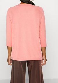 Jasnoróżowy sweter z dzianiny z rękawami o długości trzech czwartych, ze ściągaczem przy dekolcie i u dołu, zestawiony z brązowymi spodniami o szerokich nogawkach. Gładka faktura.