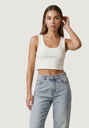 Kobieta w białym, krótkim topie i jasnoniebieskich, wysokich jeansach, stojąca na tle jednolitego jasnego tła.