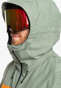 Chaqueta de esquí impermeable verde con cuello alto, detalles en negro y cordones ajustables. Incluye gafas de esquí reflectantes de gran tamaño.