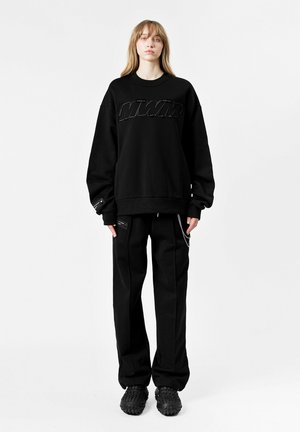 Sudadera negra de gran tamaño con el logo "MWMW" en relieve, combinada con pantalones negros que presentan cremalleras y cadenas. Un calzado texturizado completa el look.