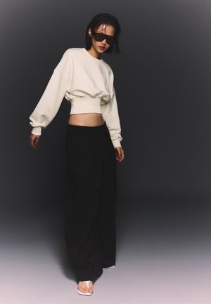 Donna che indossa una felpa bianca oversize corta, pantaloni neri larghi, grandi occhiali da sole e sandali bianchi, in piedi davanti a uno sfondo scuro.