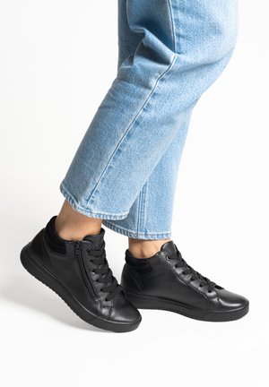 Person trägt hellblaue Jeans und schwarze Leder-High-Top-Sneaker mit Schnürsenkeln und seitlichen Reißverschlüssen, steht vor weißem Hintergrund.