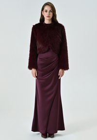 Giacca corta in pelliccia sintetica bordeaux con maniche lunghe sopra una gonna in satin drappeggiata e aderente. L'outfit presenta una texture liscia e una silhouette elegante.