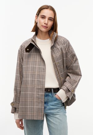 Femme portant une veste beige à carreaux par-dessus un pull blanc et un jean bleu clair, avec une main dans la poche, sur un fond uni.