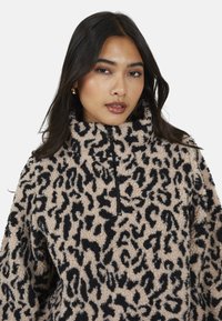 Donna con lunghi capelli scuri che indossa un pullover in pile beige e nero con stampa leopardata e colletto con zip frontale, mentre guarda verso la fotocamera.