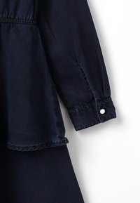 Tummansininen pitkähihoinen farkkupaita, jossa on napitettavat hihansuut ja gathers-elastinen vyötärövalinta valkoista taustaa vasten.