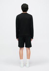 Göndör sötét hajú férfi áll egy fehér fal felé fordulva, fekete hosszú ujjú felsőt, fekete mintás rövidnadrágot, fehér zoknit és fehér tornacipőt visel.