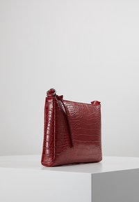 Rød taske af kunstigt krokodilleskind med en struktureret form, lynlås og kort håndtag. Tekstureret overflade med en blank finish.